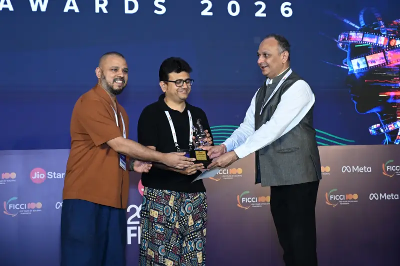 A winner accepts a FICCI BAF Awards 2026 trophy as two presenters congratulate him on stage.