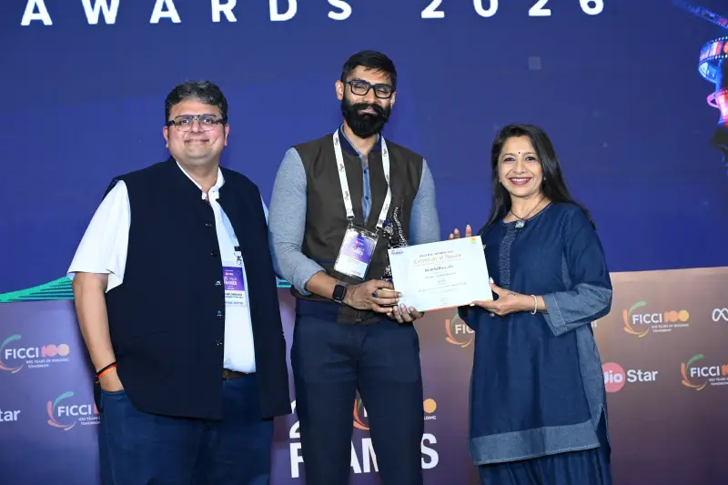A winner holds his certificate between two presenters at the FICCI BAF Awards 2026.